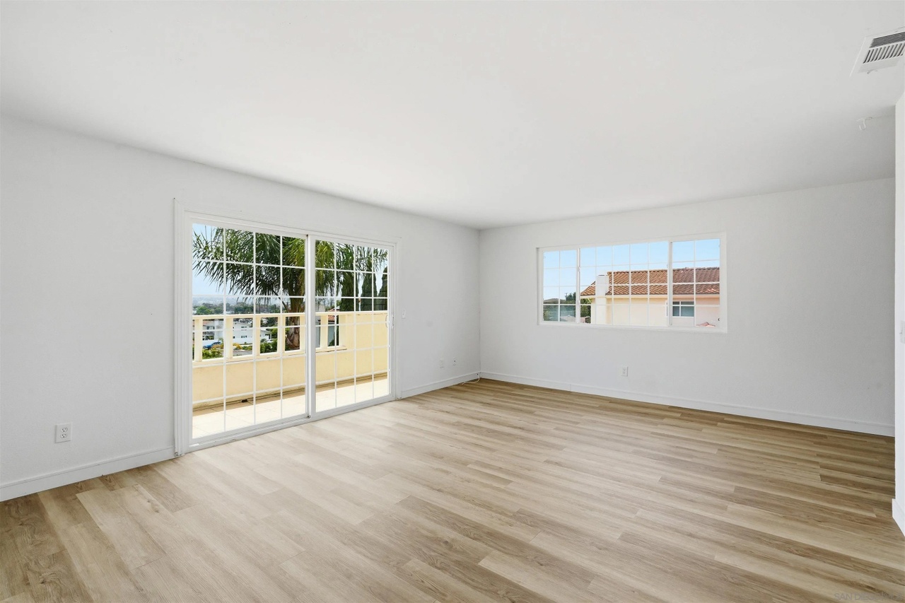 A large, empty second-level living room with new pale vinyl plank flooring, two windows, and a sliding glass door opening to a balcony