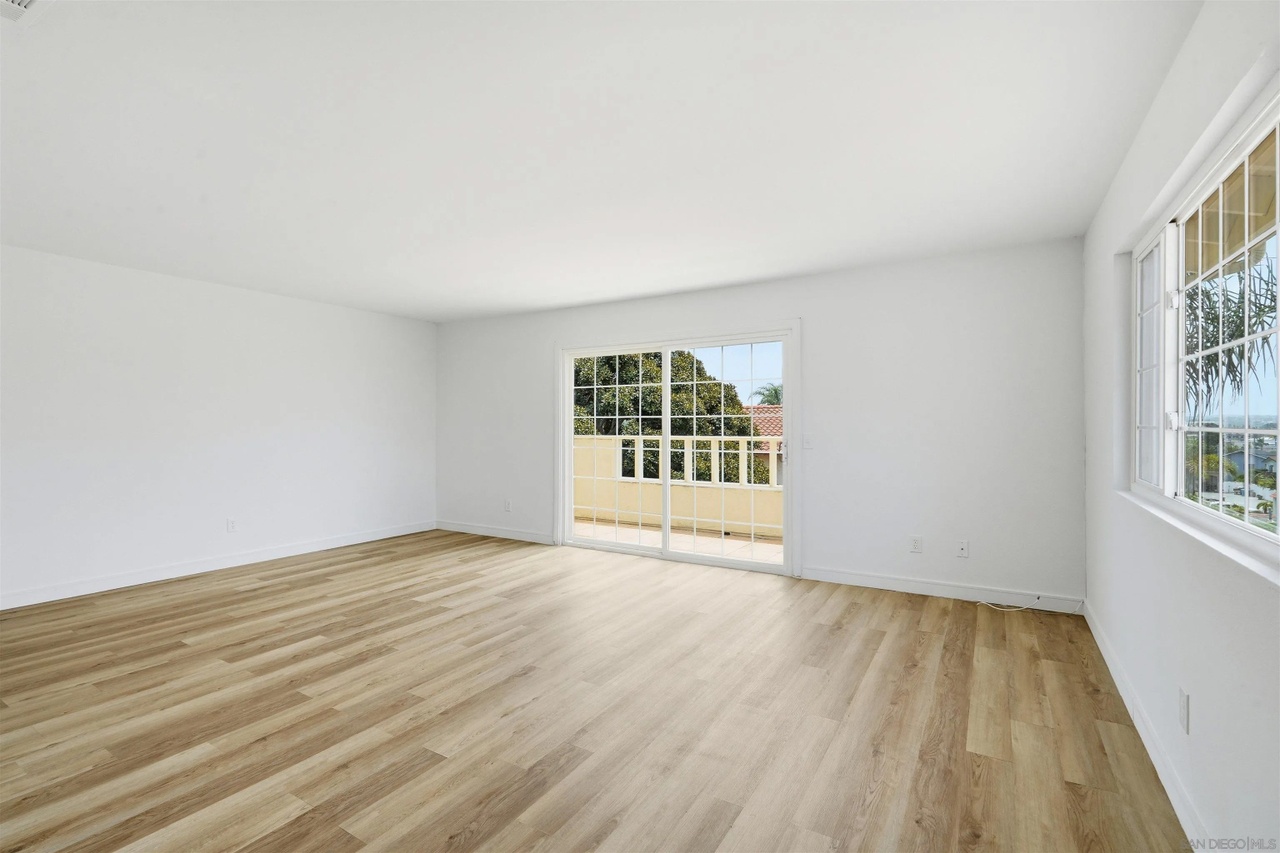A second empty living area with new vinyl plank flooring, a corner sliding glass door to the balcony, and a high window admitting bright daylight