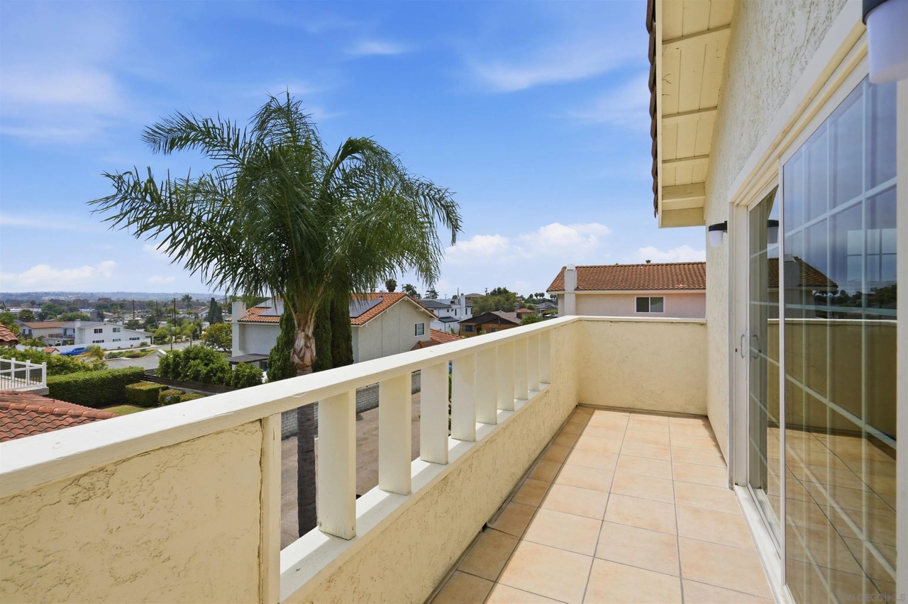 A tiled second-story balcony enclosed by a white railing, a tall palm and neighborhood rooftops in the distance under a clear sky