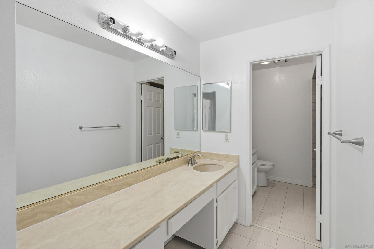 A bathroom with a long travertine vanity and single basin beneath a wide full-length mirror, with a private toilet alcove off to the side