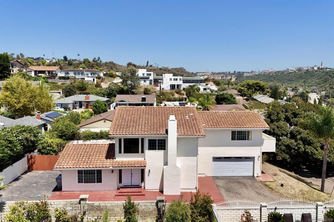 Aerial three-quarter view of 1839 Galveston Street, a two-story stucco home with a terracotta tile roof and a white two-car garage, set on a hillside above San Diego's Bay Park with canyon ridgelines behind